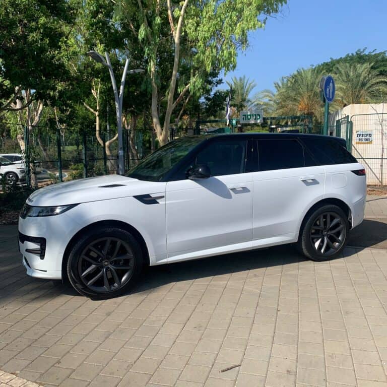 רכב SUV לבן, חלונות כהים, מבט מהצד, חניה ליד עצים וגדר.